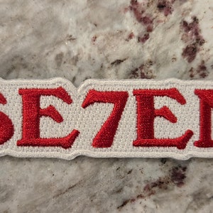 Peut inclure: Écusson en tissu blanc avec inscription brodée en rouge "SE7EN".