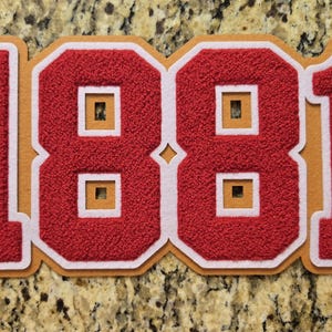 May include: A decorative sign featuring the number 1881 in red textured material with white borders, set against a gold-colored backing. The word SKEGEE is vertically embroidered in white thread within the first digit, and the number has cutouts.