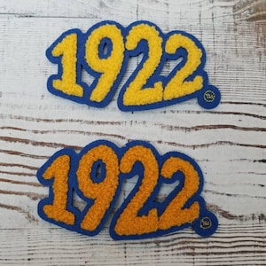 May include: Two blue felt patches with the year 1922 in yellow fuzzy letters. The patches are on a white wood background.