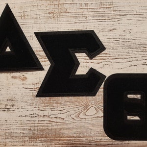 May include: Three black felt letters, a triangle, a sigma, and an omega, on a white wooden background.