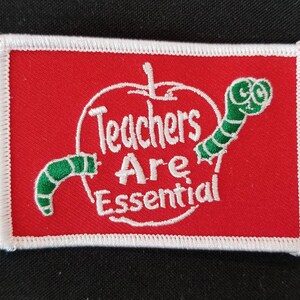 May include: Red embroidered patch with a white apple and two green worms. The apple has the text "Teachers Are Essential" inside.