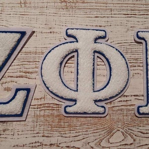 May include: Three embroidered letters: Z, Φ, and B. Each letter is white with a textured, fuzzy appearance and a blue outline. The letters are arranged on a light-colored, weathered wooden surface.
