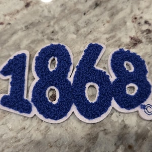 Può includere: Toppa blu e bianca con il numero "1868" in un carattere strutturato, simile alla ciniglia. I numeri sono di colore blu intenso con un bordo bianco. La toppa ha un piccolo logo circolare sul lato destro.