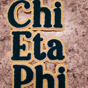 Op de afbeelding: Een close-up van een set van drie viltletterpatches. De letters zijn donkergroen met een witte omtrek en zijn genaaid op een gele vilt achtergrond. De letters spellen "Chi Eta Phi" uit.