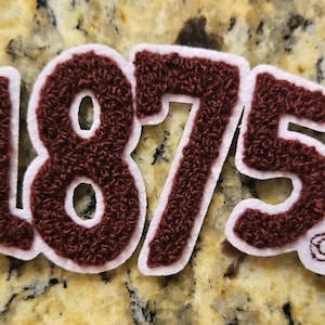 May include: A dark maroon chenille patch featuring the numbers 1875 in a bold, textured font. The numbers are outlined in a thin white border, and a small copyright symbol is visible in the bottom right corner of the number five.