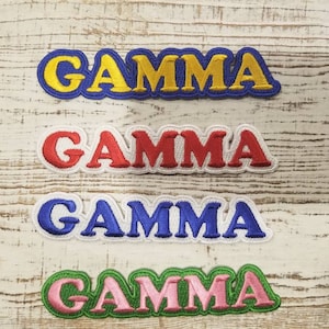 May include: Four embroidered patches with the word "GAMMA" in different colours: yellow, red, blue, and pink. The patches are on a white wooden background.