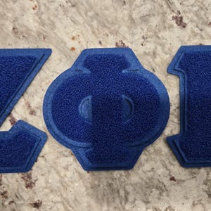May include: Three blue embroidered patches spelling out "ZOB". The letters are in a varsity style with a textured, felt-like appearance. The background is a speckled, light-colored surface.