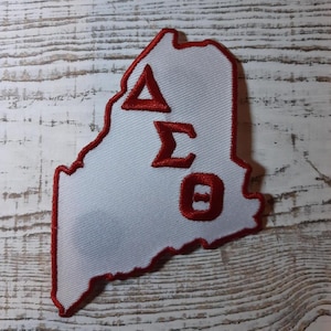 Pode incluir: Remendo de feltro branco em forma do estado do Maine com letras bordadas em vermelho que soletram "ΔΣΘ".