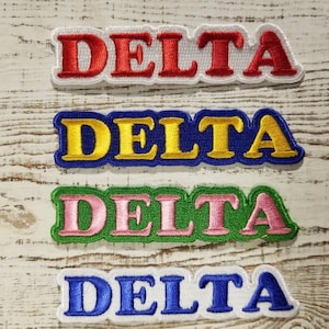 Op de afbeelding: Vier geborduurde patches met het woord "DELTA" in verschillende kleuren. De patches zijn wit met rode, blauwe, gele en groene letters.