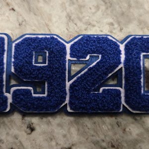 Könnte beinhalten: Ein blau-weißes Patch mit der Zahl "1920". Die Zahlen sind tiefblau mit einer flauschigen Textur und weiß umrandet. Der Aufnäher ist wahrscheinlich für eine Jacke oder andere Kleidung gedacht.