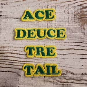 May include: Four embroidered patches with the words "ACE", "DEUCE", "TRE", and "TAIL" in green lettering with a yellow border. The patches are arranged vertically on a white wooden surface.