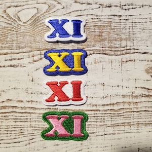 Peut inclure: Quatre patchs brodés avec le chiffre romain 'XI' dans différentes couleurs : bleu, jaune, rouge et rose. Les patchs sont sur un fond en bois blanc.