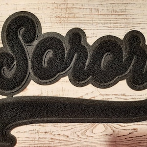 May include: Black doormat with the word "Soror" in a script font. The mat has a textured surface and a decorative border. The word "Soror" is in a stylized font, and the mat has a long, curved bottom section.