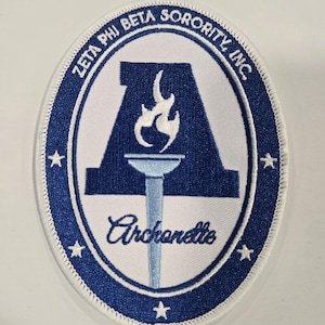 May include: An oval-shaped embroidered patch featuring a blue background with a white border. It displays the text Zeta Phi Beta Sorority, Inc. and Archonette. The design includes a large blue letter A, a light blue torch with a white flame, and stars.