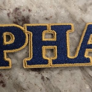 May include: A blue and gold embroidered patch with the letters "PHA" in a collegiate style font.