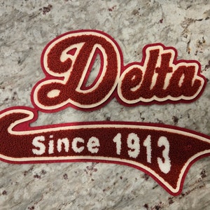 Może przedstawiać: Czerwono-biała łatka z filcu z napisem "Delta" w ozdobnym, pochylonym piśmie i tekstem "Since 1913" w białym obramowaniu.