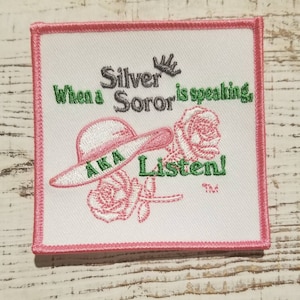 May include: White embroidered patch with pink trim. The patch features a pink rose, a white hat with the letters "AKA" on it, and the text "When a Silver Soror is speaking Listen!"