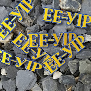 May include: Close-up of multiple yellow and blue embroidered patches with the text "EE-YIP" on a gray stone background.