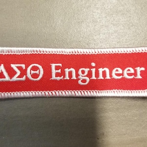 Op de afbeelding: Rode stoffen patch met witte stiksels en de tekst "ΔΣΘ Engineer" in wit.