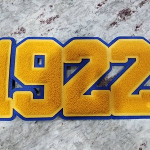 May include: A gold felt applique with blue outlining in the shape of the number 1922.