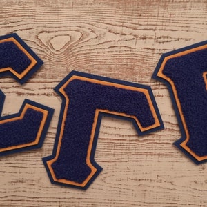 Puede incluir: Tres letras de fieltro, "E", "Γ" y "P", de color azul marino con contorno naranja. Las letras están sobre un fondo de madera clara.