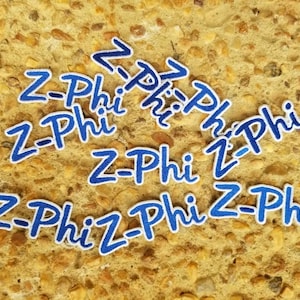 May include: A collection of blue and white embroidered patches with the text "Z-Phi" in a cursive font. The patches are scattered on a textured, light brown surface.