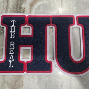 Op de afbeelding: Een marineblauwe en rode geborduurde patch met de letters "HU" en de woorden "THE REAL" verticaal aan de linkerkant. De patch heeft een witte rand, geschikt voor jassen of accessoires.