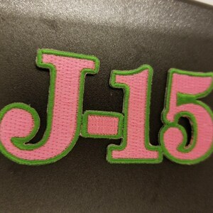May include: Pink and green felt patch with the letters "J15" in a cursive font.