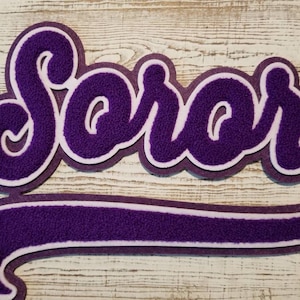 以下が含まれることがあります： 白い輪郭で「Soror」という言葉を筆記体で書いた紫色のフェルトアップリケ。アップリケには紫色のフェルトの湾曲した尾が付いています。