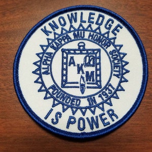 以下が含まれることがあります： 白地と青い縁取りの円形パッチ。パッチには「KNOWLEDGE IS POWER」と「ALPHA KAPPA MU HONOR SOCIETY FOUNDED IN 1937」の文字が記されています。中央のデザインには文字「A」、「K」、および「M」が含まれています。