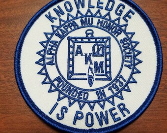May include: A circular patch with a white background and a blue border. The patch features the text "KNOWLEDGE IS POWER" and "ALPHA KAPPA MU HONOR SOCIETY FOUNDED IN 1937". The centre design includes the letters "A", "K", and "M".