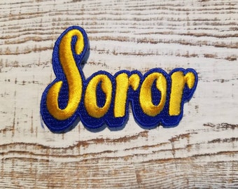 May include: A blue and gold embroidered patch with the word "Soror" in a cursive font.