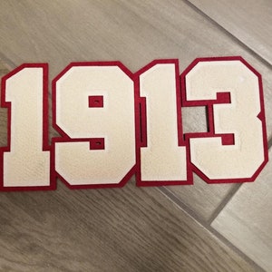Może przedstawiać: Naszywka z numerem z białego filcu z czerwonym obramowaniem. Liczby to 1913.