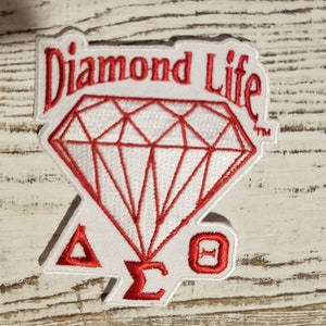 Może przedstawiać: Biała haftowana naszywka z czerwonym diamentem, napisami "Diamond Life" i literami "ΔΣΘ".