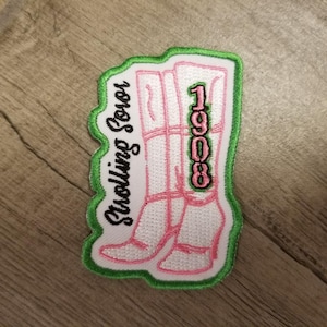 以下が含まれることがあります： 緑と白の刺繍パッチで、ピンクのブーツと「Stocking Sooer 1908」の文字が書かれています。