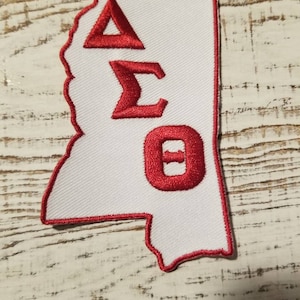 May include: A white patch with a red outline in the shape of the state of Mississippi. The letters Delta Sigma Theta are embroidered in red on the patch.