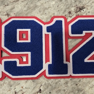 May include: A red and white felt patch with the number 1912 in blue felt.