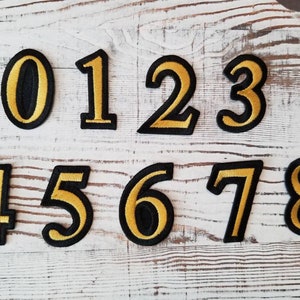 以下が含まれることがあります： 黒い輪郭と金色の数字0から8までの9つのアイロン接着パッチ。パッチは白い木製の表面にあります。