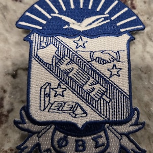 May include: Embroidered patch featuring a blue and white crest design. The crest includes a handshake, stars, and the Greek letters ΦΒΣ. The patch is bordered by a ribbon with the text "FOUNDED 1914".