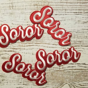 May include: Four red and white embroidered patches with the word "Soror" in a cursive font. The patches have a red border with white lettering. The patches are arranged on a white wooden surface.