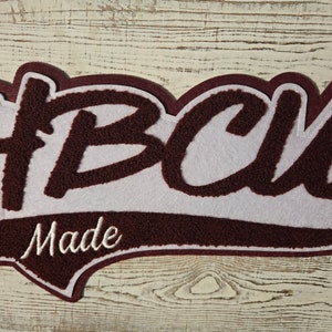 Könnte beinhalten: Ein weinrotes und weißes Patch mit dem Text "#BCU Made". Die Buchstaben "BCU" sind fett und strukturiert, mit weißem Hintergrund. Das Wort "Made" ist in Schreibschrift, ebenfalls weiß. Der Patch hat einen weinroten Rand.