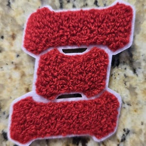May include: A red, textured patch in the shape of the letter "S" with a white border. The patch has a fuzzy, raised texture, and the color is a vibrant red. The design is simple and bold, suitable for various applications.