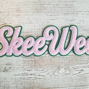 Könnte beinhalten: Ein rosa-grüner Filz-Patch mit dem Wort "Skeer Wee" in kursiver Schrift.