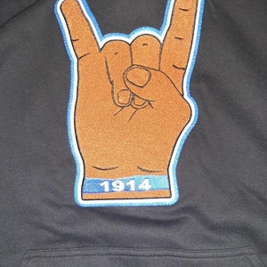 May include: A black hoodie with a brown and blue embroidered patch of a hand making the rock and roll sign. The patch has the number 1914 on it.