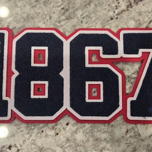 May include: A navy blue and white patch with the number "1867" in a collegiate style. The number is outlined in red and white. The word "HOWARD" is vertically embroidered in white on the left side of the number 1.