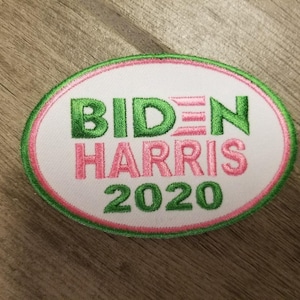 May include: Embroidered oval patch with the words "BIDEN HARRIS 2020". The text is in green and pink, with a white background and a green and pink border. The patch is on a wooden surface.