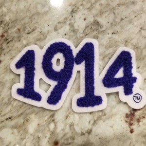 K&ouml;nnte beinhalten: Ein wei&szlig;er Filz-Patch mit der Nummer 1914 in lila, flauschigem Schriftzug. Der Patch hat ein Warenzeichen im unteren rechten Eck.