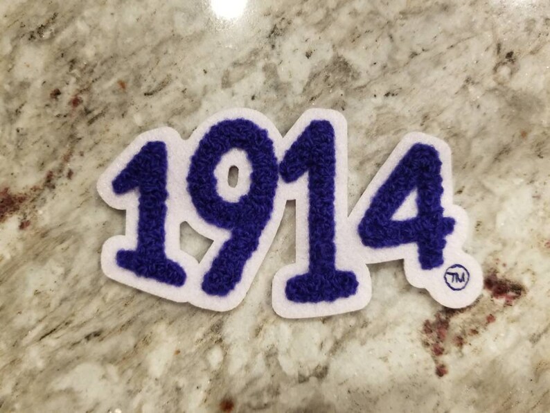 K&ouml;nnte beinhalten: Ein wei&szlig;er Filz-Patch mit der Nummer 1914 in lila, flauschigem Schriftzug. Der Patch hat ein Warenzeichen im unteren rechten Eck.