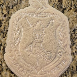 May include: A beige, textured patch featuring the Delta Sigma Theta crest. The design includes a shield with a torch, Greek letters, and a banner at the bottom with the text DELTA SIGMA THETA. The entire emblem is rendered in a monochromatic, raised, fuzzy material.