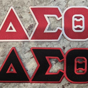 May include: Two embroidered patches featuring the Greek letters Delta, Sigma, and Omicron. One patch is red with white trim, the other is black with red trim. The patches are likely for a sorority or fraternity.